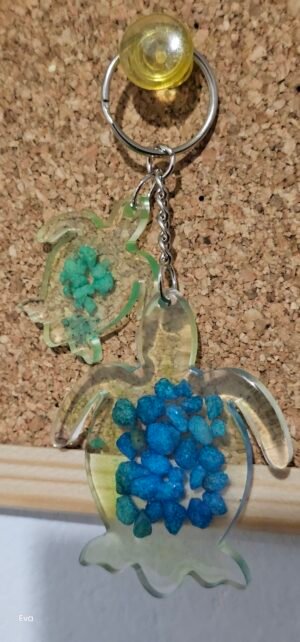 Llavero de Tortuga transparente y verde con piedras azules acompañado de una pequeña tortuga con piedras verdes.